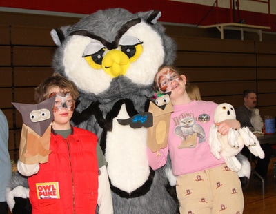 Two children hold up their owl souvenirs next to a large owl costume